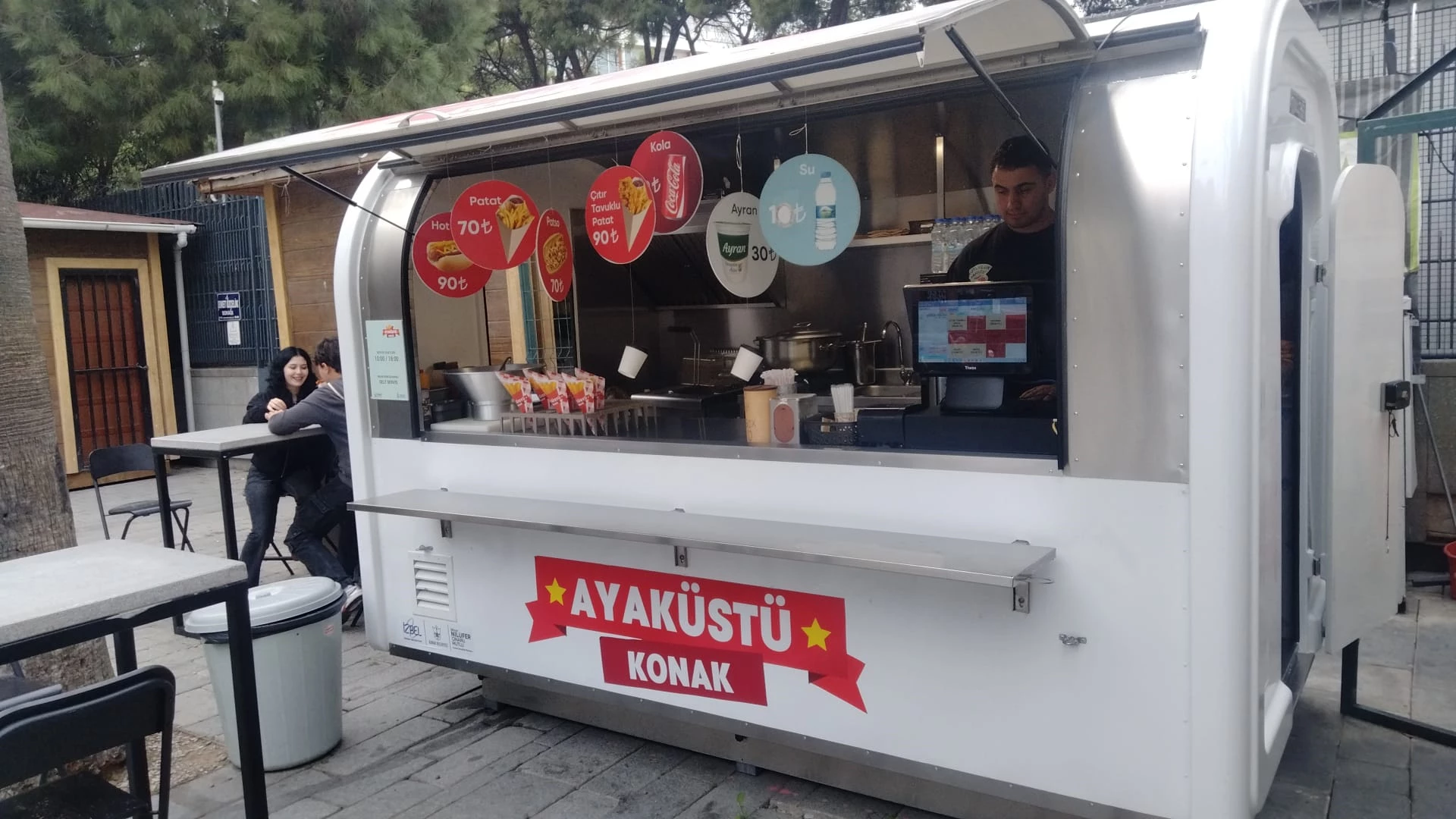 Sevgi Yolu'nda menü panoları görünen beyaz ayaküstü kiosk ve oturma alanı